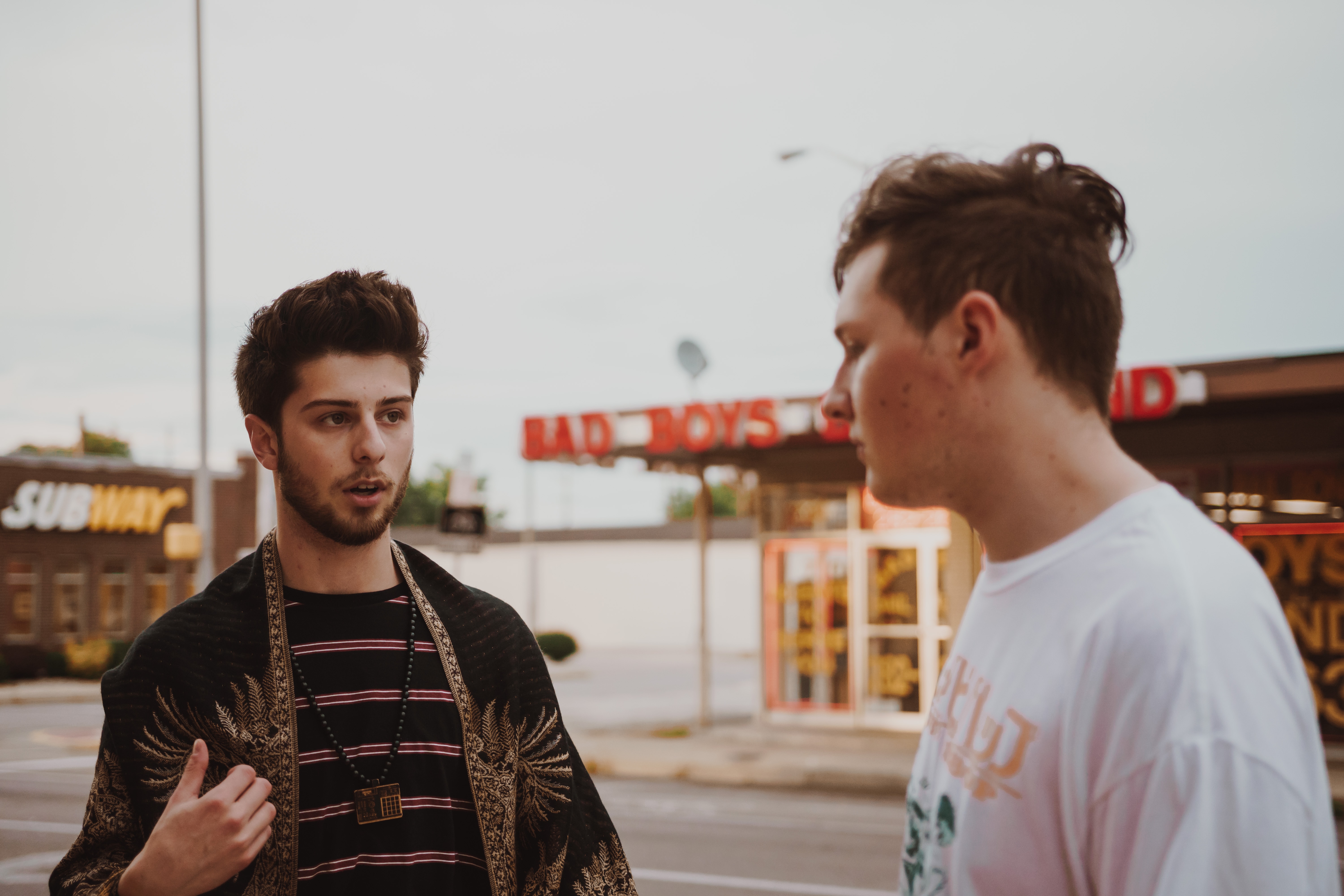 Two young men, resembling young adult cancer patients, talk on a street with a fast food restaurant and a BAD BOYS sign in the background. One gestures animatedly while the other listens, facing away from the camera.