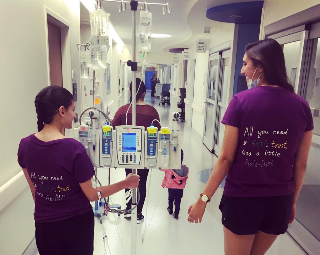 Two women in purple shirts with IV poles walk down a hospital hallway with a child and another adult. The shirts read, “All you need is faith, trust, and a little pixie dust.”.