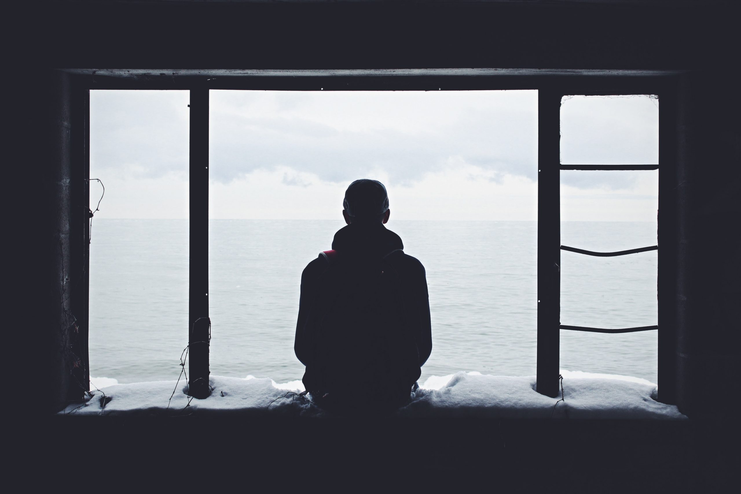 A person sits on a ledge inside a window frame, facing a calm, overcast sea. Snow covers the windowsill, and a metal ladder is attached to the right. The quiet scene invites thoughts of how to support someone in isolation.
