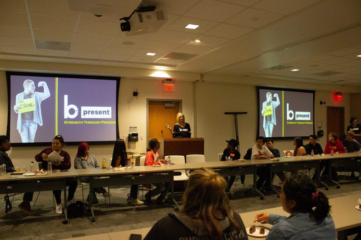 A presenter stands at a podium in a conference room, addressing a seated audience. Two screens display a slide about young adult cancer and an image of a person holding a sign that says IM STRONG.