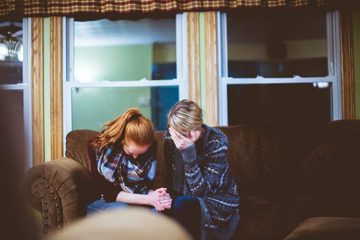 Two women sit closely together on a couch, heads bowed and hands clasped, using gentle talking tips to comfort each other in a moment of sadness or distress in a warmly lit living room.