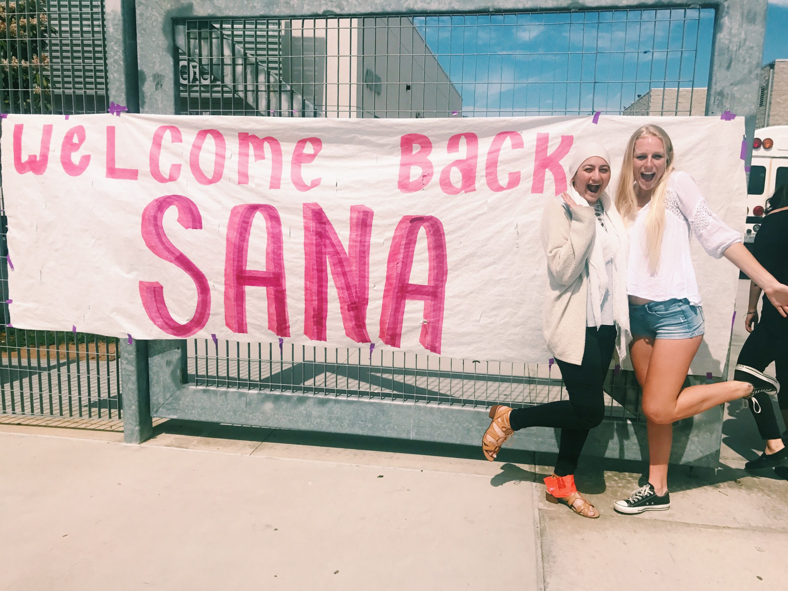 Two young women happily pose in front of a large handwritten banner that says Welcome Back Sana in pink letters, welcoming their classmate outside on a sunny day.