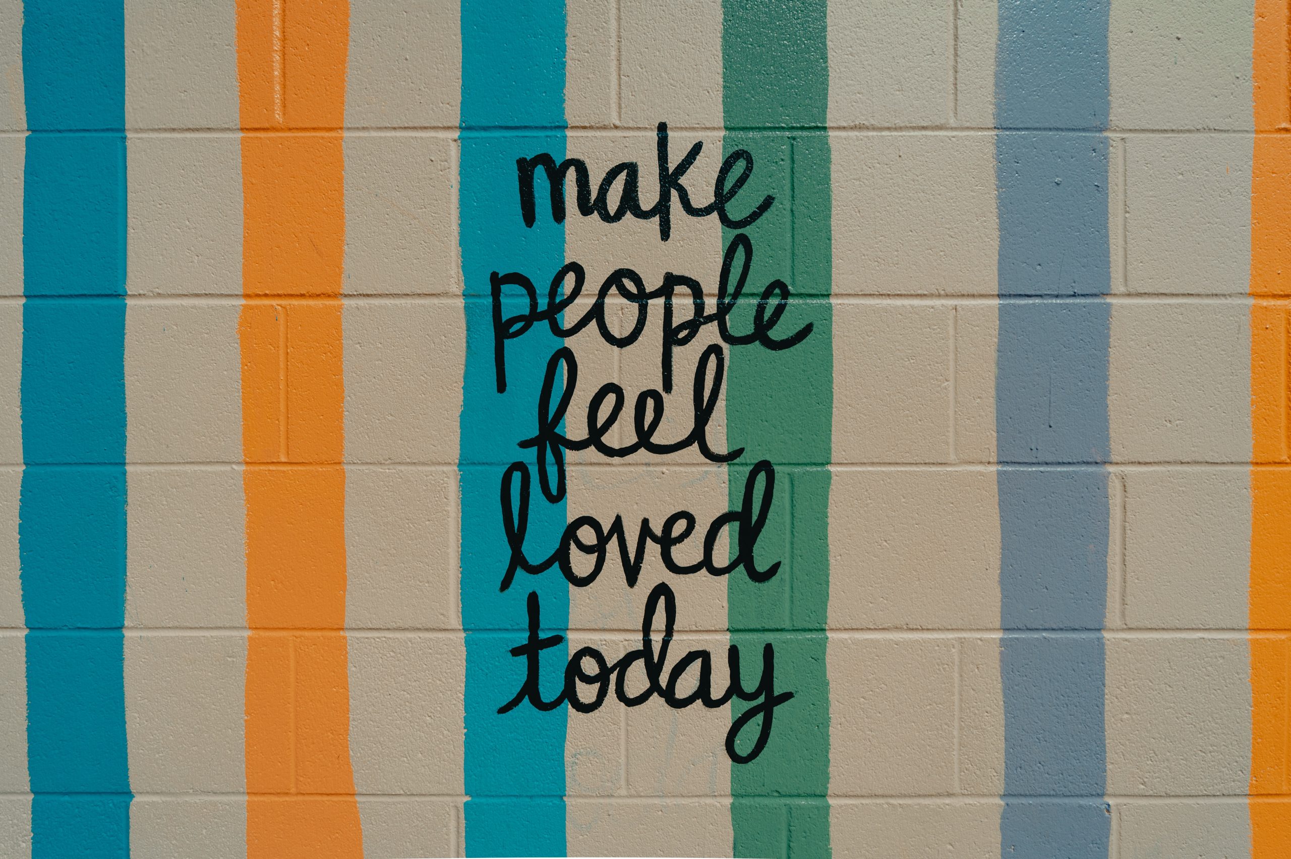A painted wall features vertical stripes in blue, orange, green, and beige. In the center, black handwritten text reads, make people feel loved today—an uplifting reminder to show empathy.