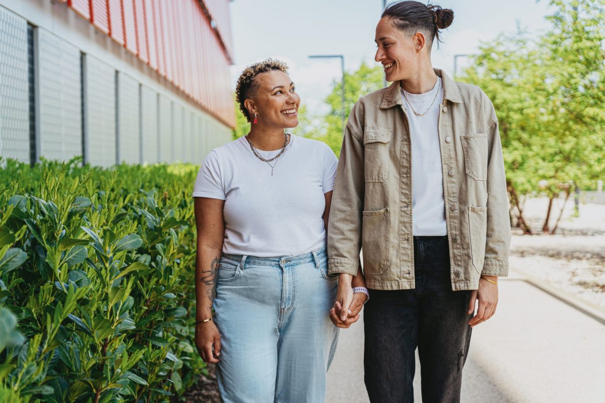 Two romantic partners holding hands and smiling at each other while walking outside on a sunny day, surrounded by green bushes and trees.