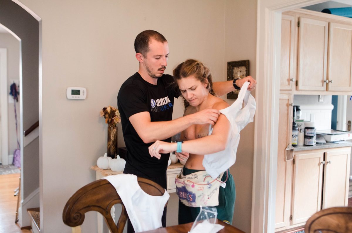 A man helps a woman put on a white shirt in a home kitchen, fostering an environment of normalcy. The woman, wearing a medical device around her waist, looks uncomfortable, while the scene conveys support and caregiving.