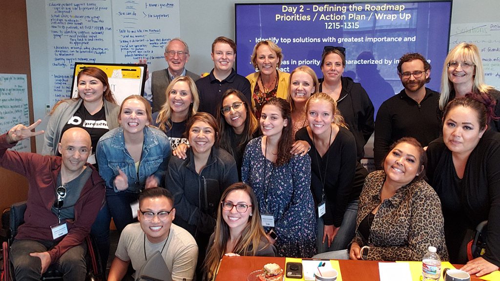A group of people pose and smile together in a conference room. Behind them, a screen displays a workshop agenda, and large sheets of handwritten notes are on the walls. Everyone appears friendly and enthusiastic.