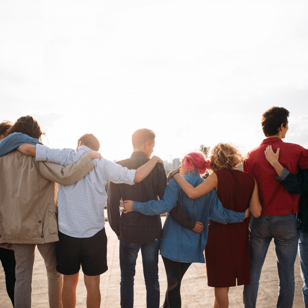 A group of seven people, representing the b-present Foundation, stands outdoors with their backs to the camera, arms around each other’s shoulders and facing the bright sky. The group appears friendly and supportive.