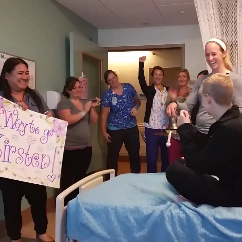 A group of smiling people stand around a hospital bed, celebrating. One person holds a decorated sign that says Way to go, Kirsten! while another helps a child sitting on the bed ring a bell. Everyone looks happy and supportive.