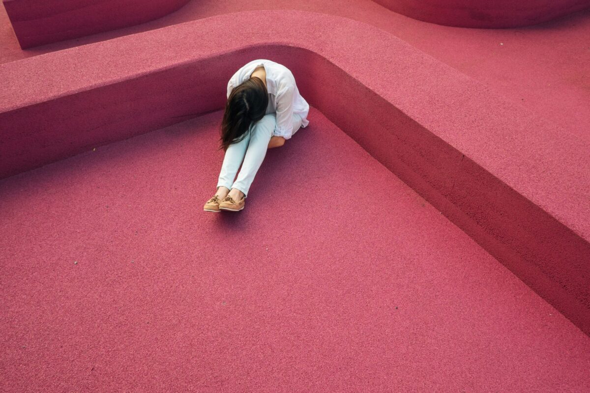 A person in light clothing sits on a bright pink surface with their head down and knees drawn up, surrounded by curved pink walls, creating a striking geometric scene that evokes the emotional weight of cancer ghosting.
