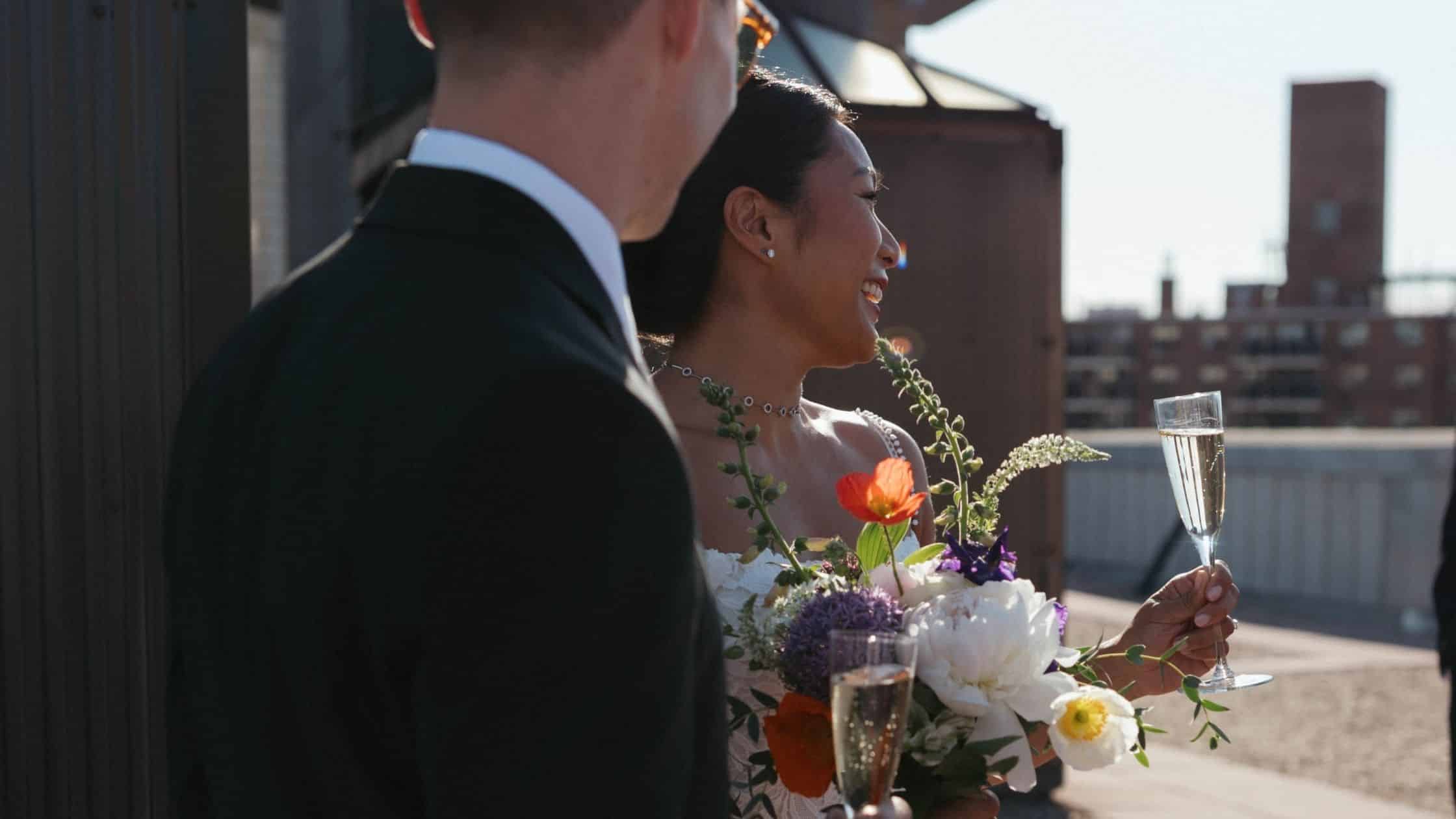 A bride holding a bouquet and a glass of champagne smiles beside a groom in a suit on a sunny rooftop with city buildings in the background, celebrating their perfect day after months of thoughtful wedding planning.