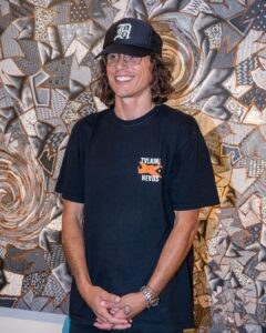 A smiling person with long hair, glasses, and a black cap stands in front of a colorful, abstract patterned artwork. They wear a black Tylking Hevds t-shirt—perfect for Pop Culture Picks—and have their hands clasped in front.