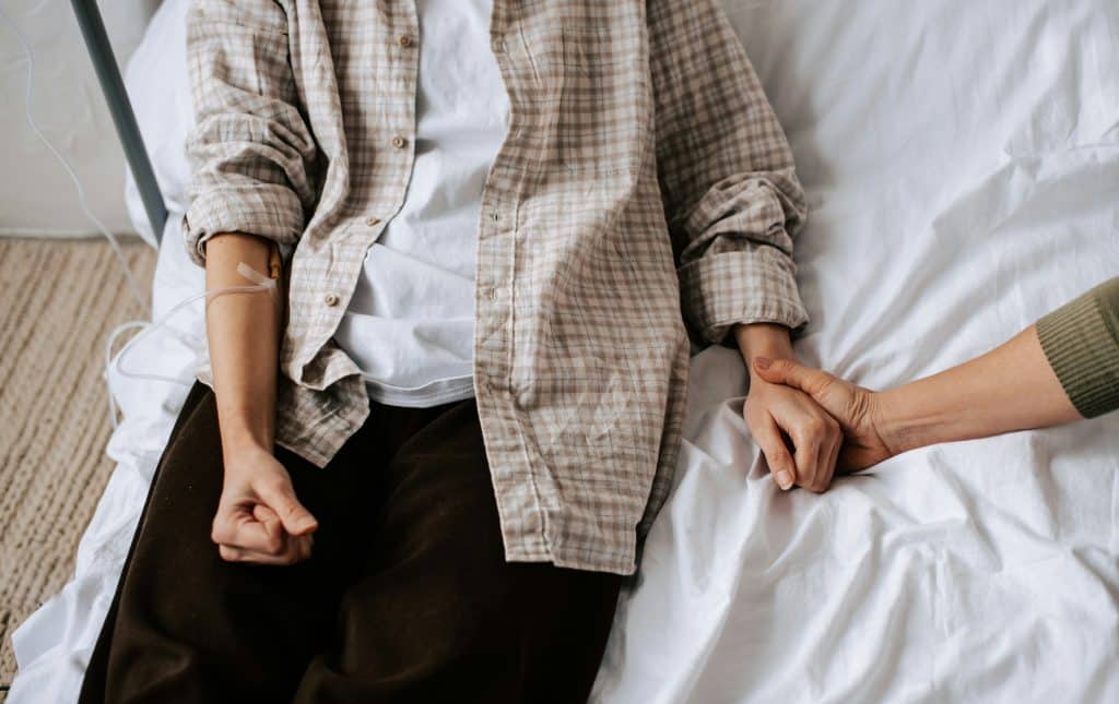 A person lies in bed with an IV in their arm, while another gently holds their hand, offering comfort and support during a moment of grief.