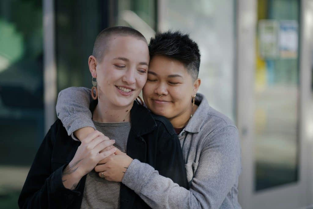 Two people stand close together outdoors, one hugging the other from behind. Both are smiling with closed eyes, showing affection and comfort—capturing the supportive bond often found among AYA Cancer survivors. Blurred glass doors and light fill the background.
