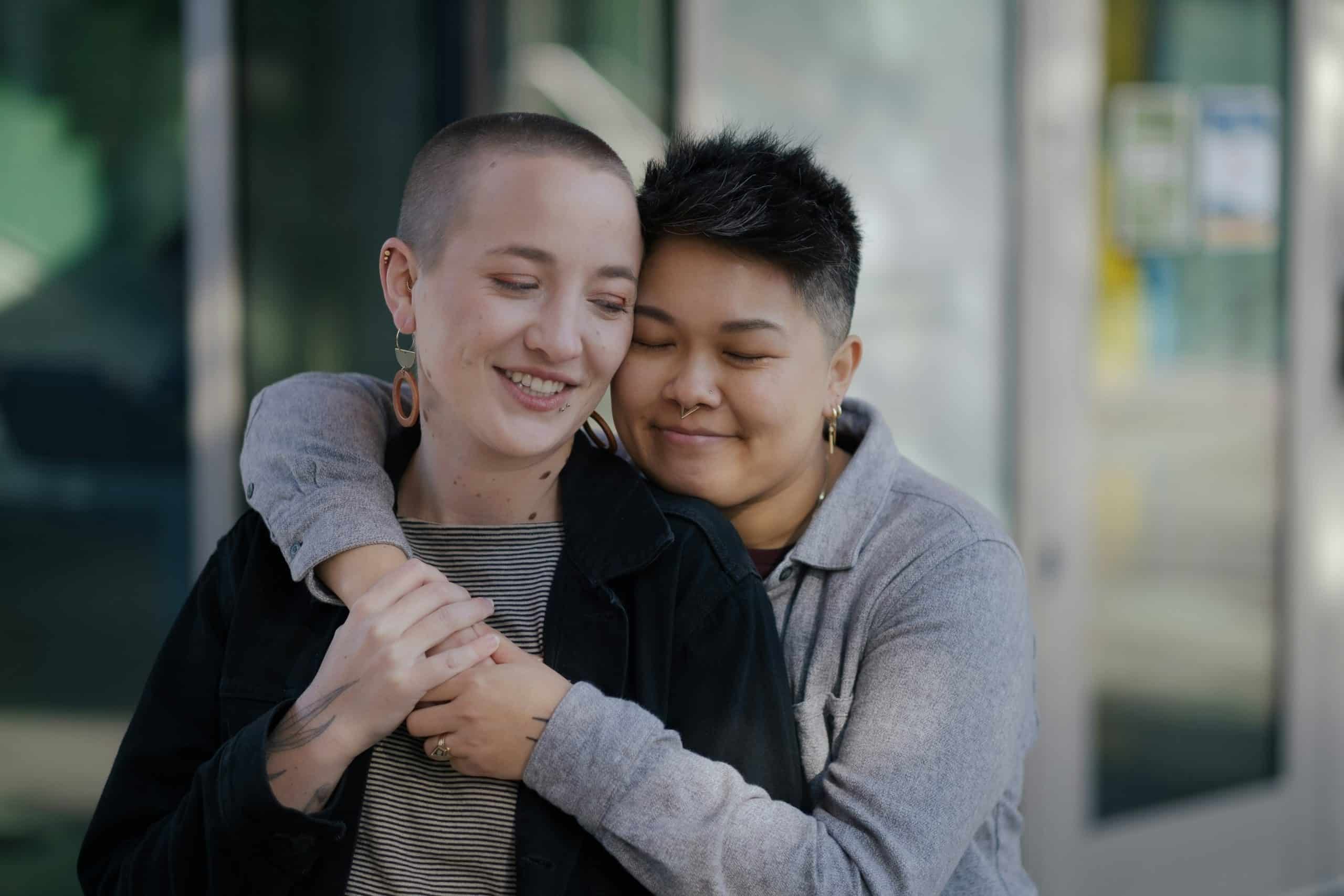 Two people stand close together outdoors, one hugging the other from behind. Both are smiling with closed eyes, showing affection and comfort—capturing the supportive bond often found among AYA Cancer survivors. Blurred glass doors and light fill the background.