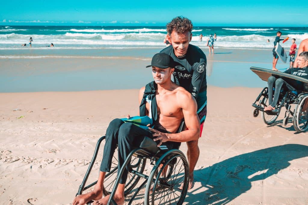 A man in a wheelchair sits shirtless on a sandy beach, holding a surfboard, while his caregiver stands behind him, smiling. Waves and other beachgoers are visible in the background.
