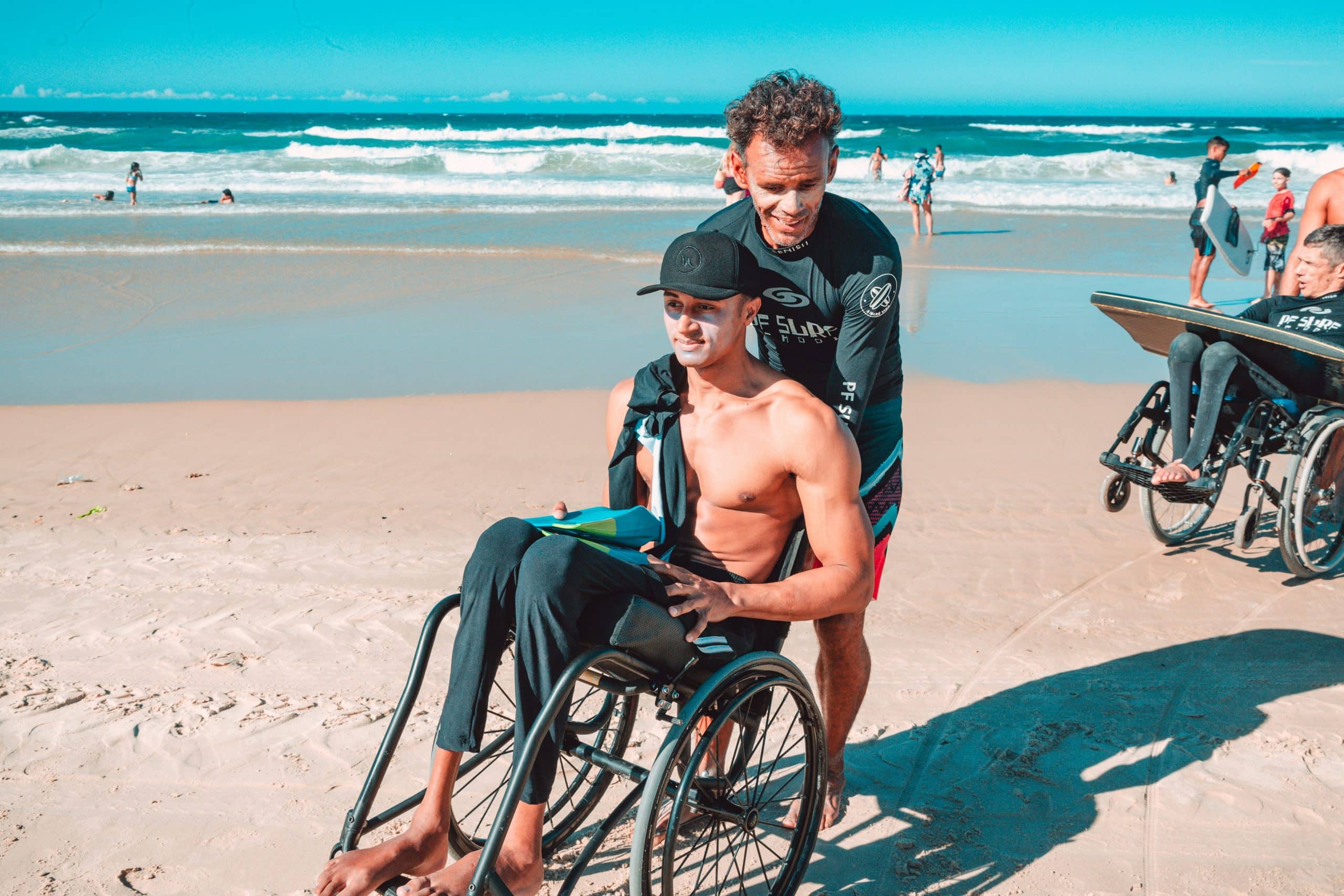 A man in a wheelchair sits shirtless on a sandy beach, holding a surfboard, while his caregiver stands behind him, smiling. Waves and other beachgoers are visible in the background.