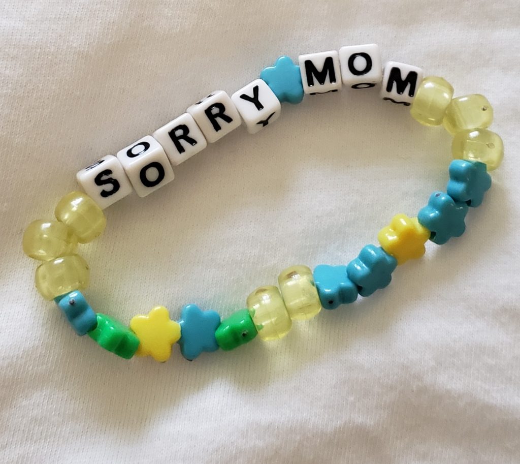 A beaded EDC bracelet on a white background spells SORRY MOM with white square letter beads, decorated with blue, yellow, and green beads in fun shapes like stars and flowers.