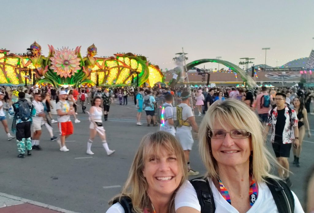 Two women smiling and taking a selfie at an outdoor festival, surrounded by colorful, illuminated dragon and flower decorations. The lively crowd, reminiscent of the vibrant EDC atmosphere, includes many people in festive or bright clothing.