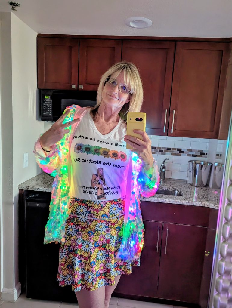 A woman stands in a kitchen taking a mirror selfie, dressed for EDC in a glowing, multicolored LED jacket over a white T-shirt with text, a floral skirt, and glasses, while flashing a peace sign.