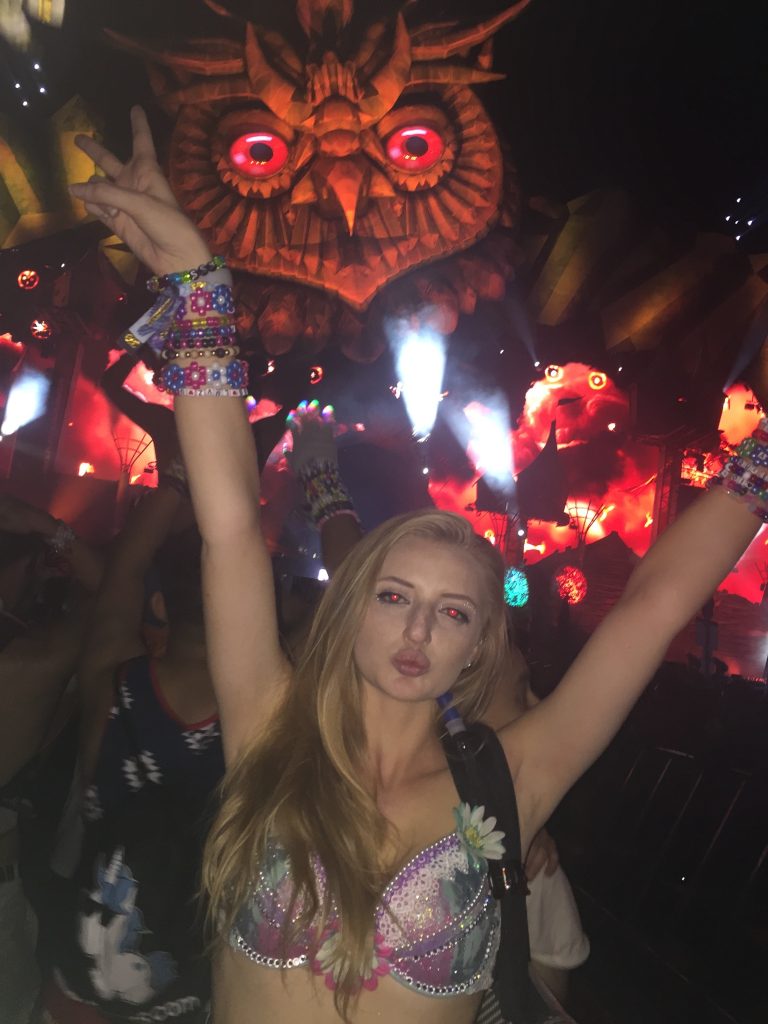 A woman in a decorated bra top and colorful beaded bracelets poses with arms raised at EDC, a nighttime festival. Bright lights and a large illuminated owl figure glow in the background.