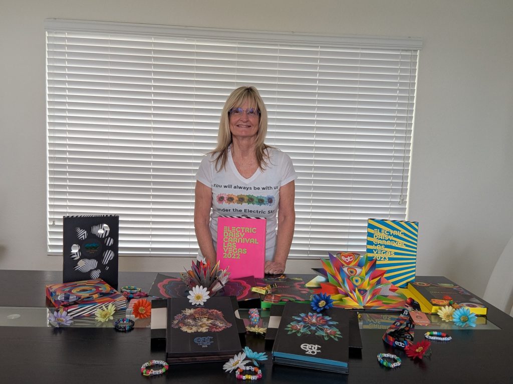 A woman with blond hair and glasses stands smiling behind a table displaying colorful pop-up books, EDC bracelets, flowers, and art. She wears a white T-shirt with text, and there are large blinds in the background.