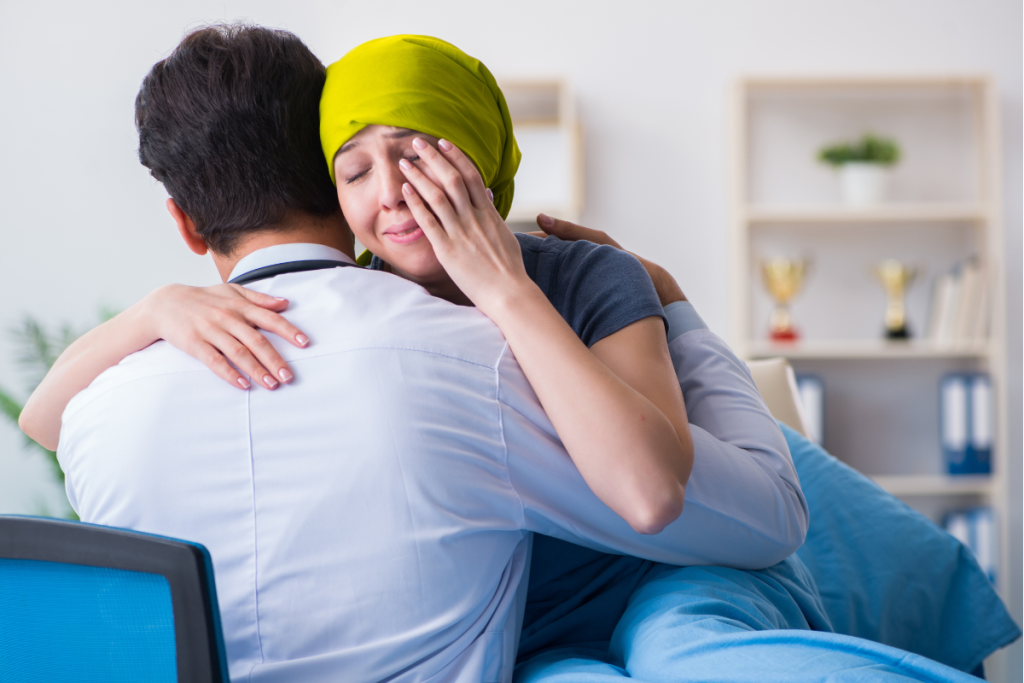 A doctor embraces a woman in a green headscarf, who appears emotional and is wiping tears while sitting on a hospital bed, offering comfort and support as she faces setbacks in a medical setting.