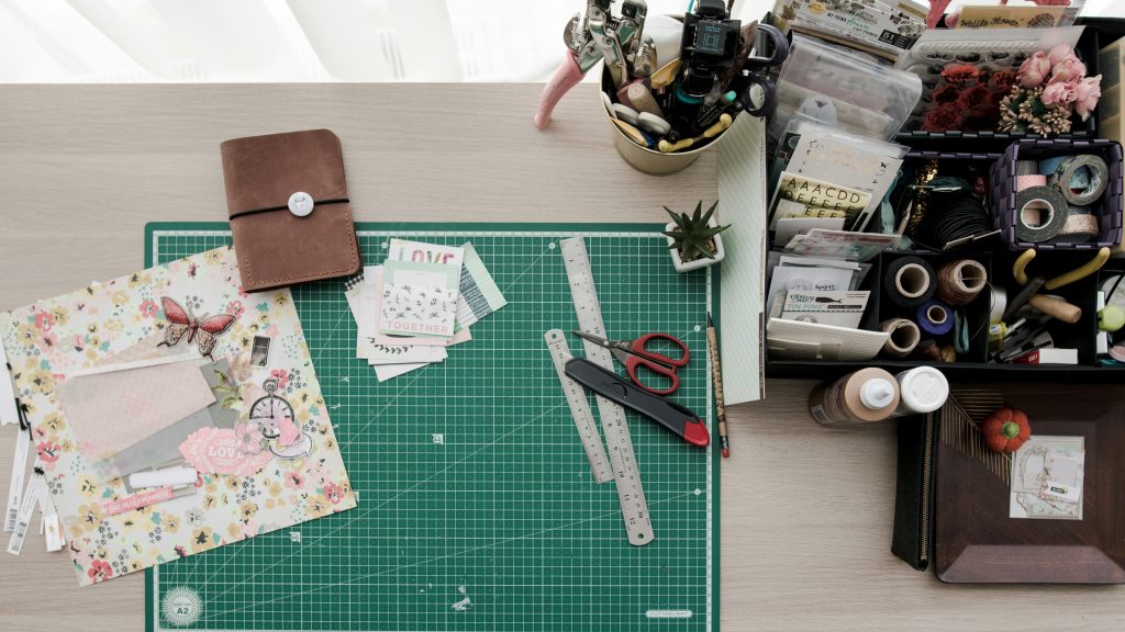 A tidy workspace with a green cutting mat, scrapbook paper, scissors, rulers, a brown notebook, and organized stationery—perfect for creating a vision board and arranging craft supplies on a light wood desk.