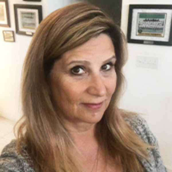 A woman with long, light brown hair and a gray top looks at the camera, smiling slightly. Framed pictures and certificates hang on the white wall behind her.