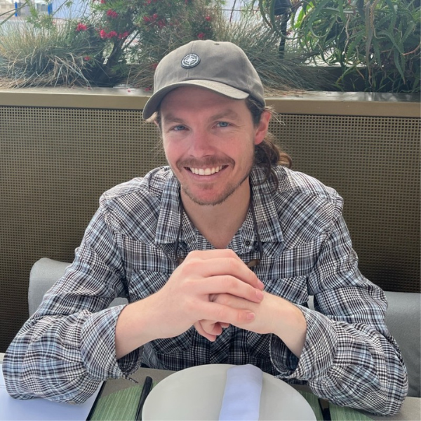 A smiling man with long hair, wearing a gray cap and plaid shirt, sits at a table set with a plate and napkin. There are plants and flowers in the background.