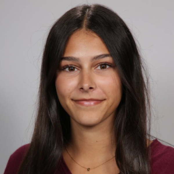A young woman with long, straight dark hair smiles softly at the camera. She is wearing a maroon top and a delicate necklace, with a plain light gray background behind her.