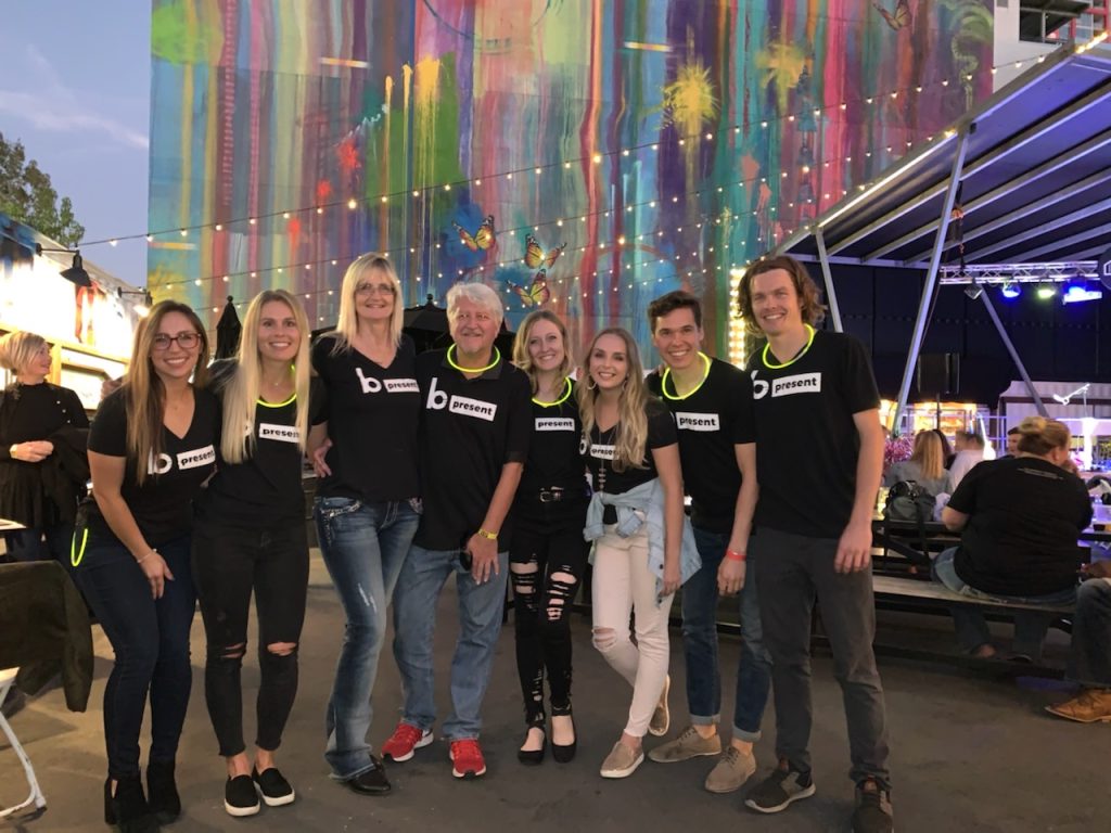 Eight people stand closely together smiling at an outdoor event, all wearing black b present t-shirts and glow necklaces—celebrating support for young adult cancer fighters amid colorful lights and butterfly decorations.