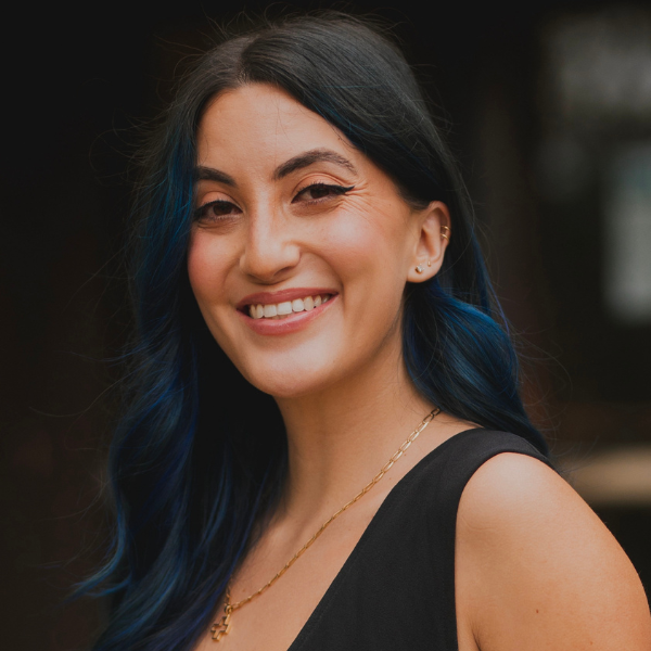 A woman with long dark hair and blue highlights smiles at the camera. She is wearing a sleeveless black top and a gold necklace. The background is blurred and dark.