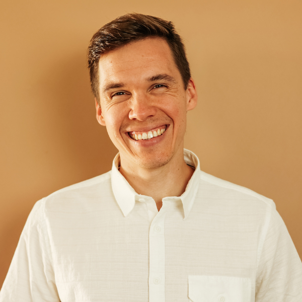A man with short brown hair, wearing a white button-up shirt, smiles warmly while standing in front of a plain beige background.