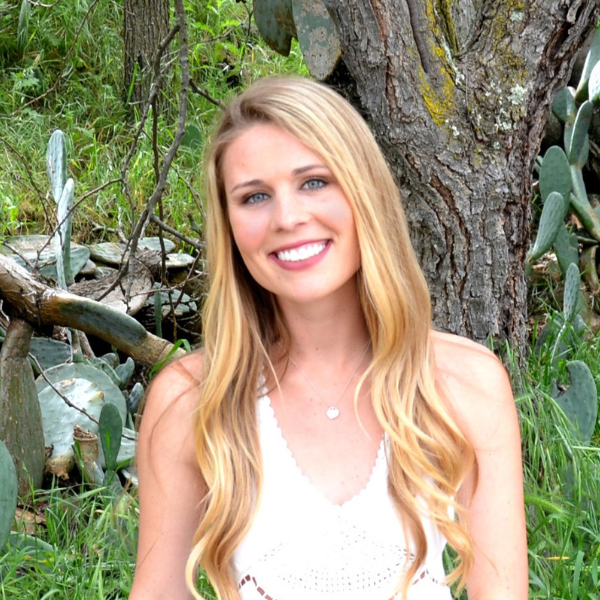 A smiling woman with long blonde hair in a white sleeveless top sits outdoors in front of a tree and green plants, including prickly pear cactus.