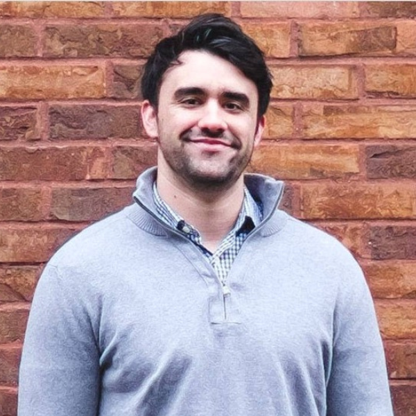 A man with dark hair and a short beard is smiling at the camera. He is wearing a light gray pullover sweater over a checkered shirt, standing in front of a red brick wall.