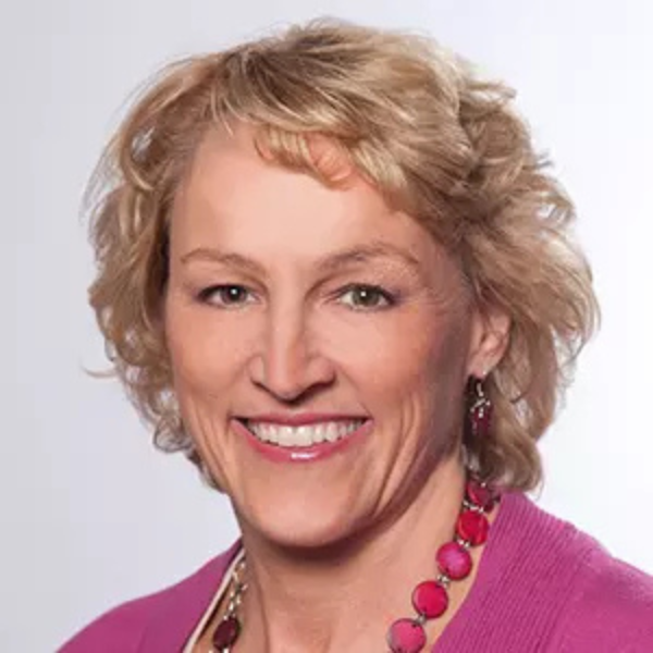 A woman with short, curly blonde hair smiles at the camera. She is wearing a pink sweater, a pink beaded necklace, and matching earrings. The background is plain and light-colored.
