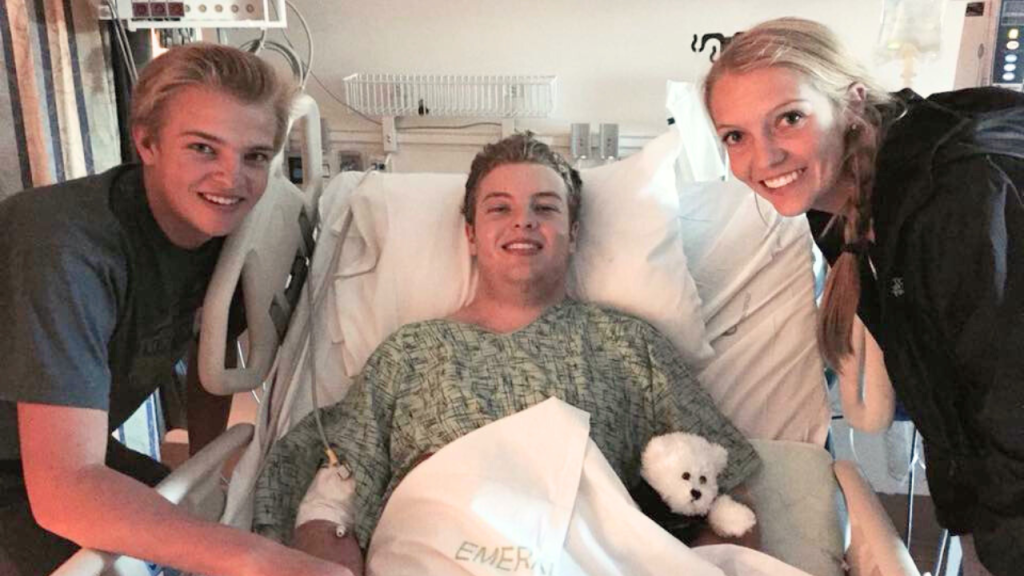A young person lies in a hospital bed smiling, holding a small stuffed animal, while two people stand on either side of the bed and smile at the camera. Medical equipment is visible in the background.