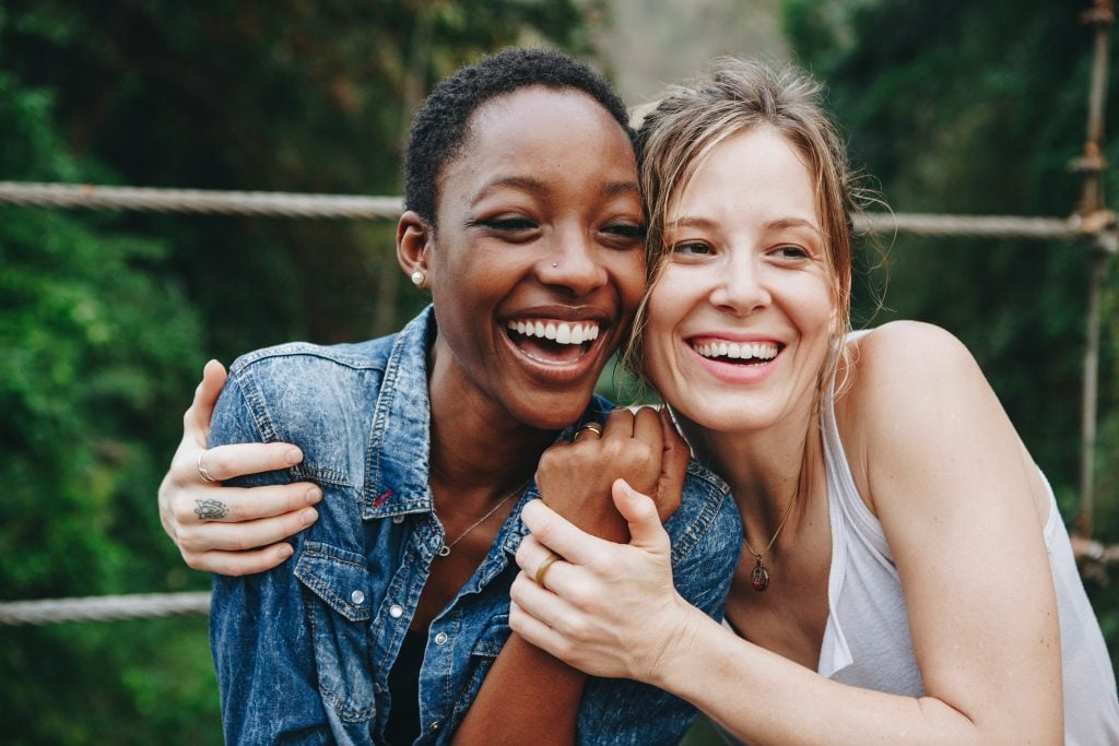 Two women smiling and hugging each other outdoors, standing in front of a rope bridge with green trees in the background. Both appear happy and relaxed, enjoying each others company.