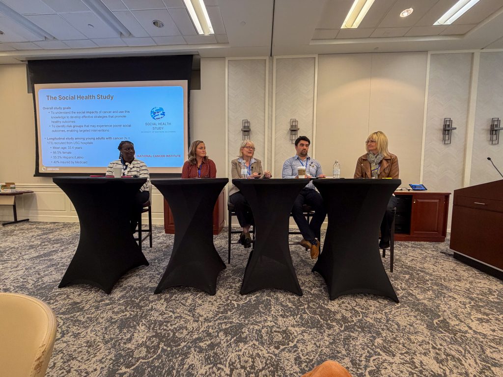 Five people sit at a panel table in a conference room, discussing The Social Health Study as shown on a projector screen beside them. Each has a microphone and water bottle, with audience chairs in the foreground.