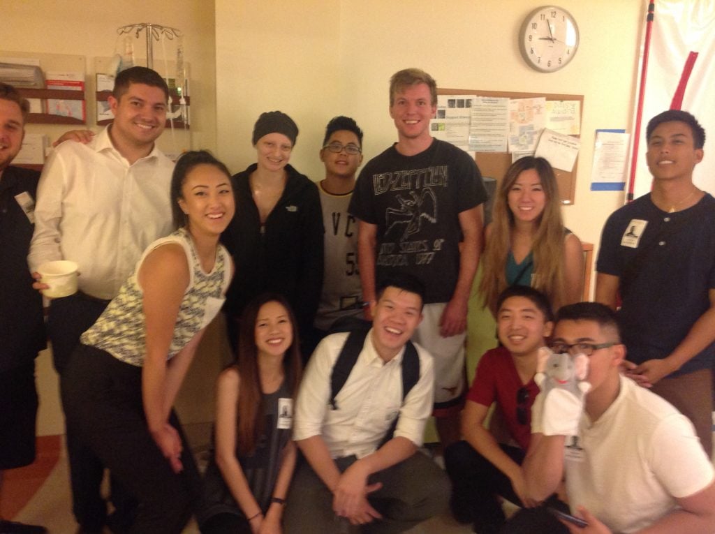 A group of smiling young adults poses together in a hospital room; some are standing, some kneeling, and one wears a hospital wristband and head covering—like those featured in a young adult cancer guide. A clock and medical equipment are visible in the background.