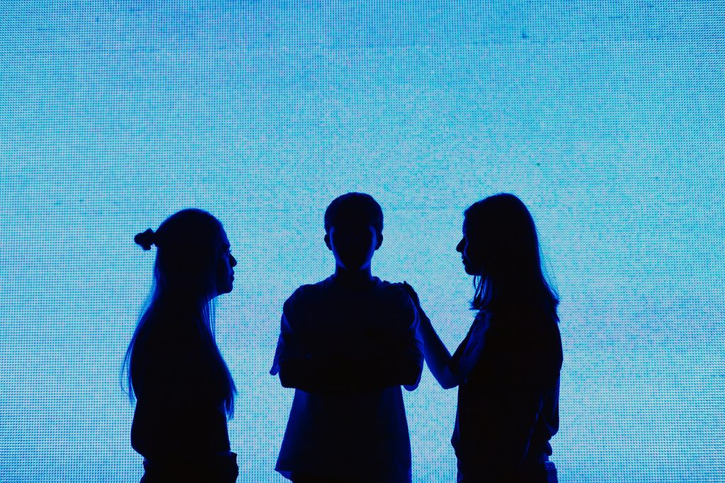 Three people are standing in front of a bright blue background. They appear as dark silhouettes, with two facing each other and one standing between them with crossed arms, evoking the tension of an unexpected cancer diagnosis.